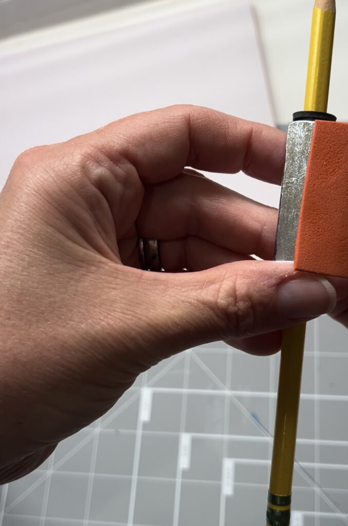 A hand wrapping a strip of colorful sticky-back craft foam around the metal nuts on a pencil to create a soft, comfortable grip.
