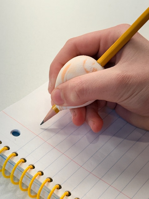 Side view of the built up foam pencil grip using an orange and white beauty blender. A child's hand write on a white, spiral bound piece of paper using the handwriting aid.